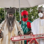 "Emir of kano wearing Kokowar Alkyabba attire holding a stick, standing on chariot under royal umbrella"