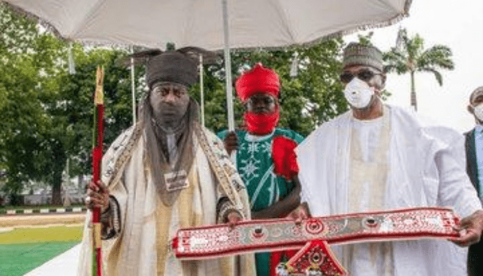 "Emir of kano wearing Kokowar Alkyabba attire holding a stick, standing on chariot under royal umbrella"