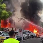 fire from an attack consumes cars while fire fighters try to put it off