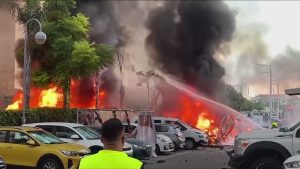 fire from an attack consumes cars while fire fighters try to put it off
