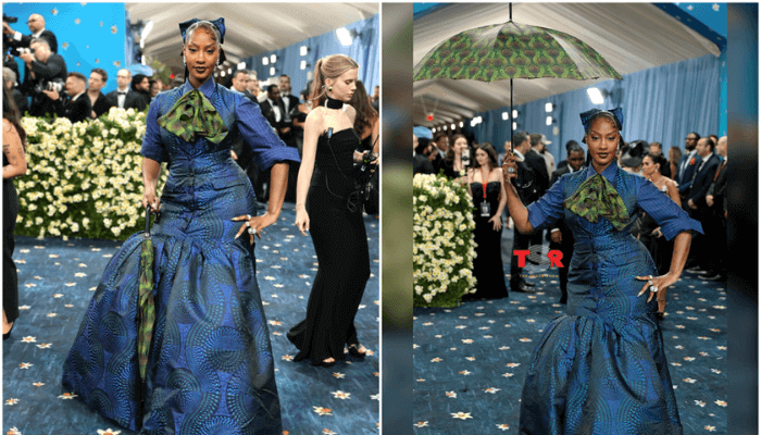 Tems wearing a buttoned-up suit dress made with blue Ankara Fabric, with a green cravat around her neck and a matching green umbrella.
